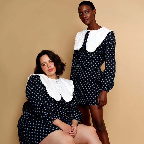 Duas modelos vestindo a mesma roupa da C&A: um vestido preto com estampa de bolinhas brancas e uma gola branca com babados. A primeira é uma menina branca e gorda, com cabelo liso e curto, e a outra é negra e magra, de cabeço raspado.