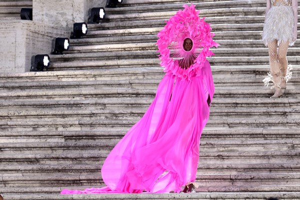 Modelo negra e jovem, com cabelo curto raspado, desfilando em escadas em uma praça de Roma, na Itália. É uma apresentação da marca Valentino e ela usa um vestido rosa e um adereço escultural na cabeça na mesma cor.