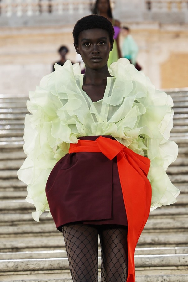 Modelo negra e jovem, com cabelo curto raspado, desfilando em escadas em uma praça de Roma, na Itália. É uma apresentação da marca Valentino e ela usa uma blusa de babado verde neon em tule, uma saia mini na cor vinho com um laço laranja amarrado e uma meia calça arrastão.