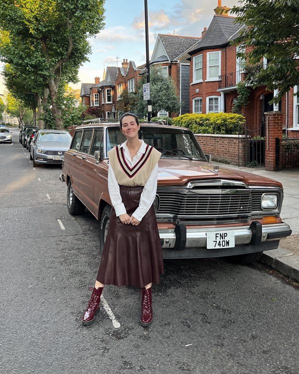 Uma mulher branca e jovem, de cabelos castanhos amarrados, posando para foto encostada em um carro antigo marrom. Ela veste uma camisa branca de botão, um colete marrom por cima, uma calça longa de couro plissada e uma bota estilo coturno vinho