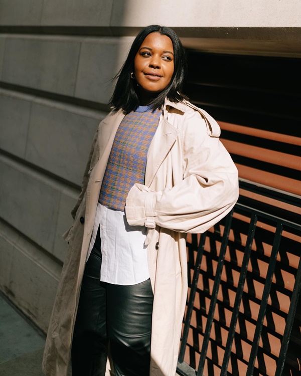 Mulher jovem e negra, de cabelos lisos médios, posando para foto na rua, em frente a um muro cinza. Ela veste uma camisa de botão branca, um colete cinza por cima, calça preta de couro e um sobretudo bege.