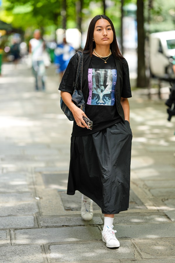 Mulher asiática e jovem andando pelas ruas de Paris. Ela tem os cabelos pretos lisos e cumpridos e veste: uma camiseta preta, uma sala longa preta, tênis branco da Adidas com meias longas.