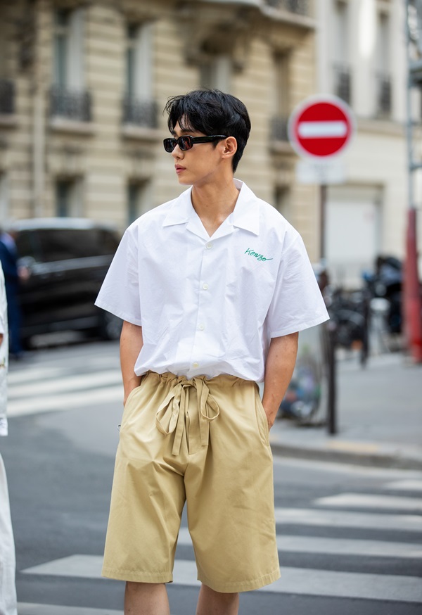 Homem branco asiático, com cabelos curtos e castanho, posando para foto nas ruas de Paris. Ele usa uma camisa de botão branca de manga curta, uma bermuda bege e óculos escuros.