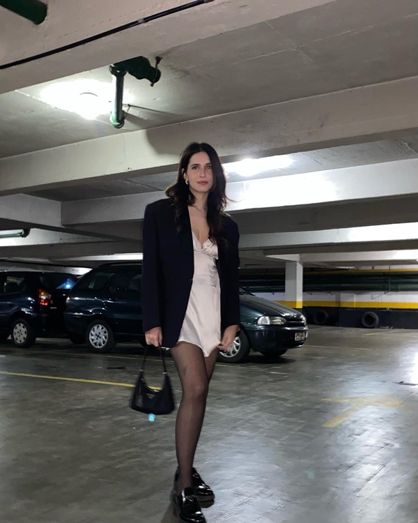 Mulher branca, jovem e de cabelos lisos longos, posando para foto em um estacionamento. Ela usa um vestido de cetim branco, meia calça preta, sapato mocassim preto e blazer também preto.