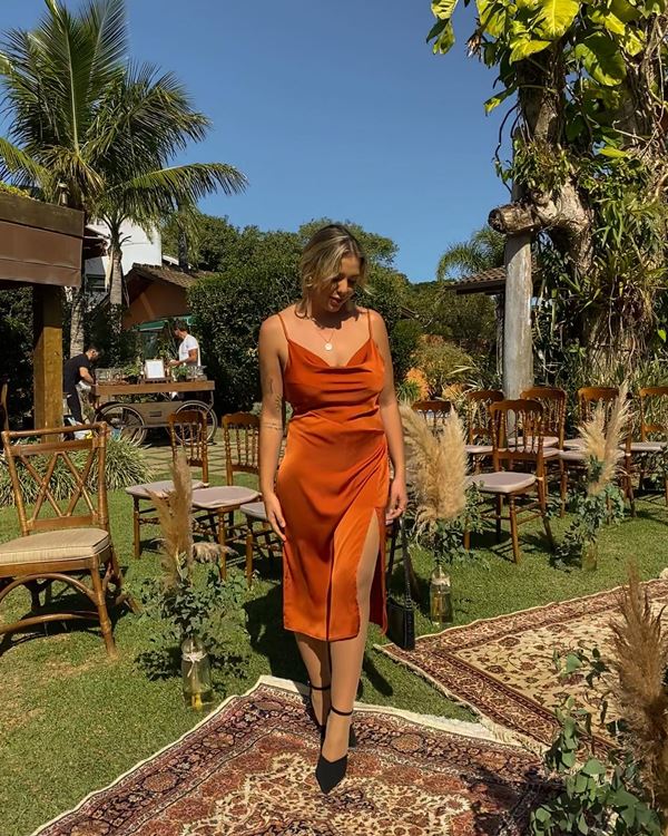 Mulher jovem, branca e loira posando para foto ao ar libre em uma estrutura que parece um salão de festas. Ela usa um vestido laranja cobreado de alcinha e rente ao corpo e uma sandália de salto preta.