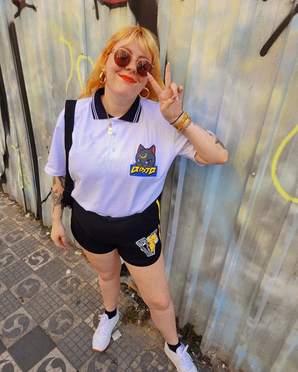 Mulher branca e jovem, com cabelo loiro, liso e longo posando para foto em um tapume prata. Ela usa uma camisa polo branca, um shorts pretos, tênis branco e óculos escuros.