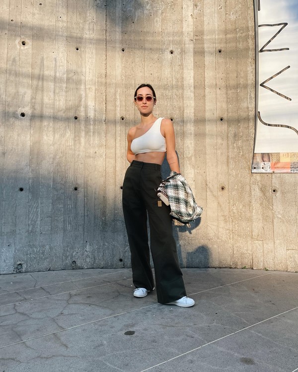 Mulher branca e jovem, com cabelo loiro, liso e presto posando para foto em uma estrutura cinza de concreto. Ela usa um top de uma alça só branco, que deixa a barriga à mostra, calça folgada preta, tênis branco e, nas mãos, segura uma jaqueta xadrez preto e branco.