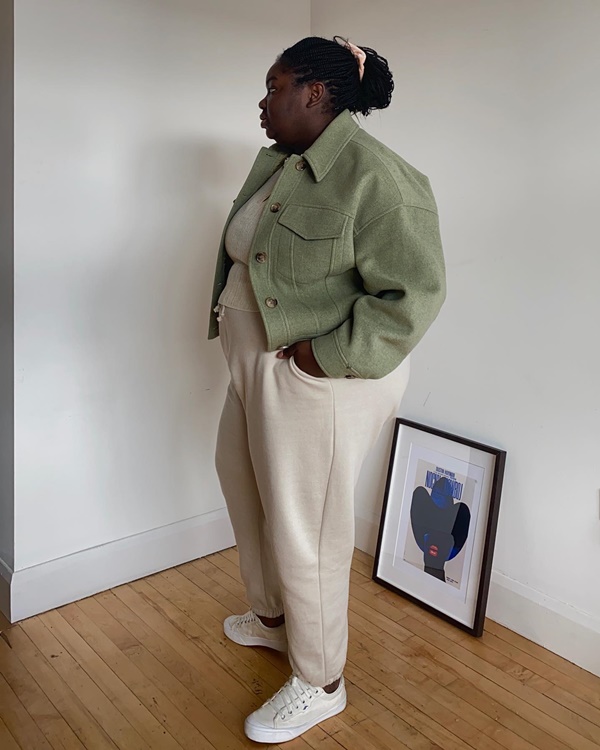 Mulher negra e jovem, com cabelos longos trançados, posando para foto na sala de casa. Ela usa uma blusa e calça de moletom bege, um casaco verde militar e um tênis branco.