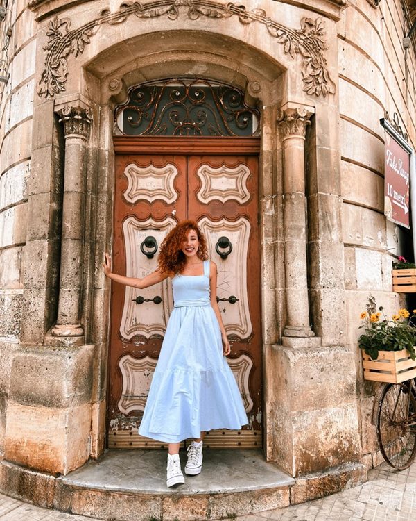 Mulher branca e jovem, com cabelos cacheados e ruivos, posando para foto na porta de uma rua em Roma. Ela usa um top e uma saia midi azul clara e um tênis branco All Star