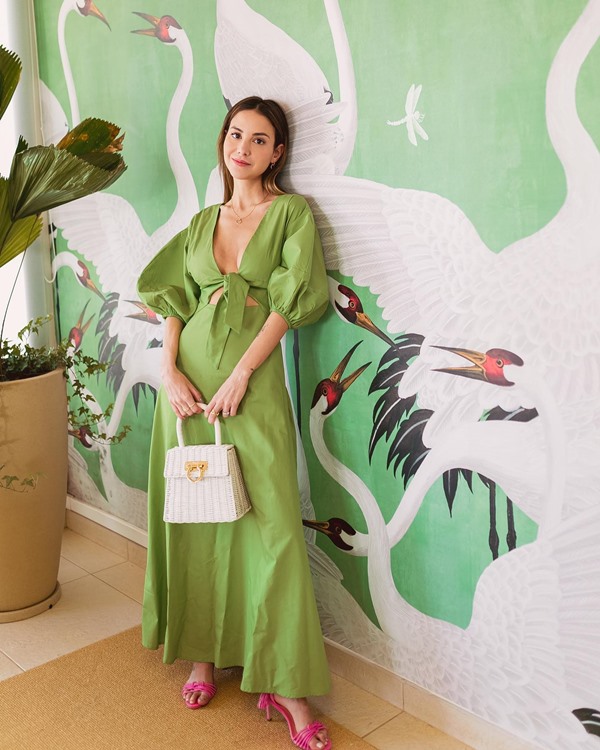 Mulher branca e jovem, com cabelo liso e louro, posando para foto na sala de casa. Ela usa um vestido longo verde, com mangas bufantes, e uma bolsa de palha branca