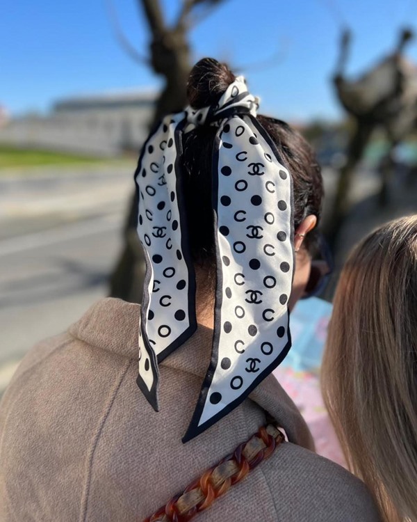 Mulher branca e jovem mostrando um penteado no cabelo com lenço. Ela usa um casaco de inverno bege e, para fazer o penteado, um lenço de fita preto e branco