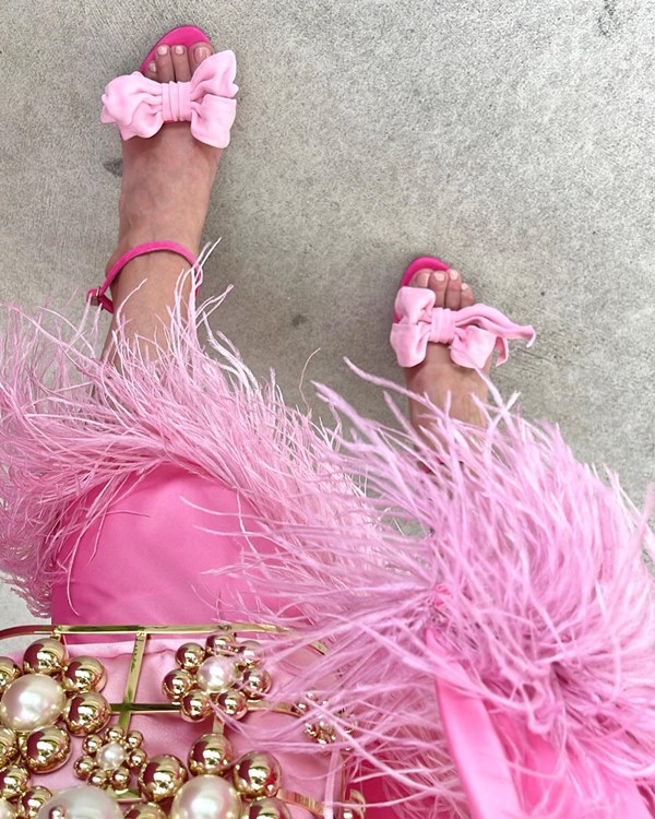 Foto de detalhes do look de uma blogueira de moda. Na foto é possível ver que ela usa um vestido rosa com plumas, uma bolsa dourada com pérolas e uma sandália rosa com um laço em um tom mais rosa de claro