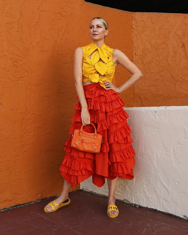 Mulher branca e jovem, com cabelo loiro amarrado em um coque, posando para foto em um fundo marrom. Ela usa um top amarelo com laços e uma saia laranja com babados. 