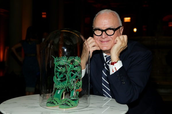 O estilista Manolo Blahnik, um homem branco e idoso, sentado com os braços em uma mesa branca de mármore ao lado de um sapato desenhado por ele, na cor verde e de salto alto. Ele usa óculos de grau com armação escura, um terno preto e uma gravata listrada preta e branca