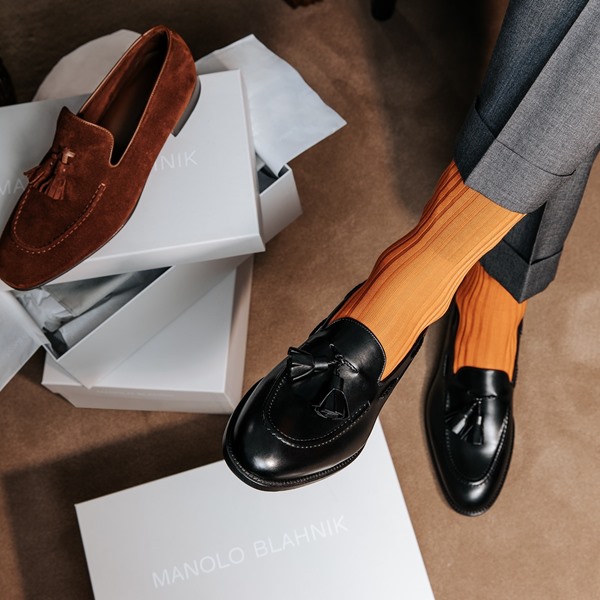 Sapato estilo mocassim, em couro preto, da marca Manolo Blahnik. A pessoa que veste o calçado usa também uma meia laranja e uma calça cinza. Ao lado do seu pé é possível ver uma pilha de caixas da marca e outro mocassim, do mesmo modelo, mas em couro marrom