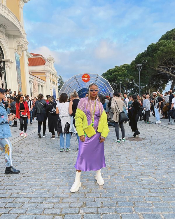 Mulher jovem e negra, com cabelos trançados longos e loiros, posando para foto no meio de uma rua lotada de pessoas. Ela usa uma blusa de manga longa e um vestido de cetim longo, ambos lilás, e um casaco verde neon por cima. Nos pés, calça um tênis branco com meias cumpridas também brancas.