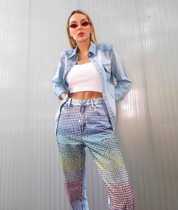 Mulher jovem e branca, com cabelos lisos loiros, posando para foto em uma parede de concreto em uma área ao ar livre. Ela usa um top branco, camisa jeans por cima, calça jeans com aplicações de strass coloridos e um óculos de sol com lentes vermelhas.