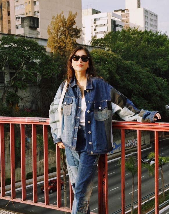 Mulher jovem e branca, com cabelos lisos castanhos, posando para foto no bairro da Liberdade, em São Paulo. Ela usa uma camiseta branca e um conjunto de jaqueta e calça jeans de retalhos do tecido em diferentes lavagens