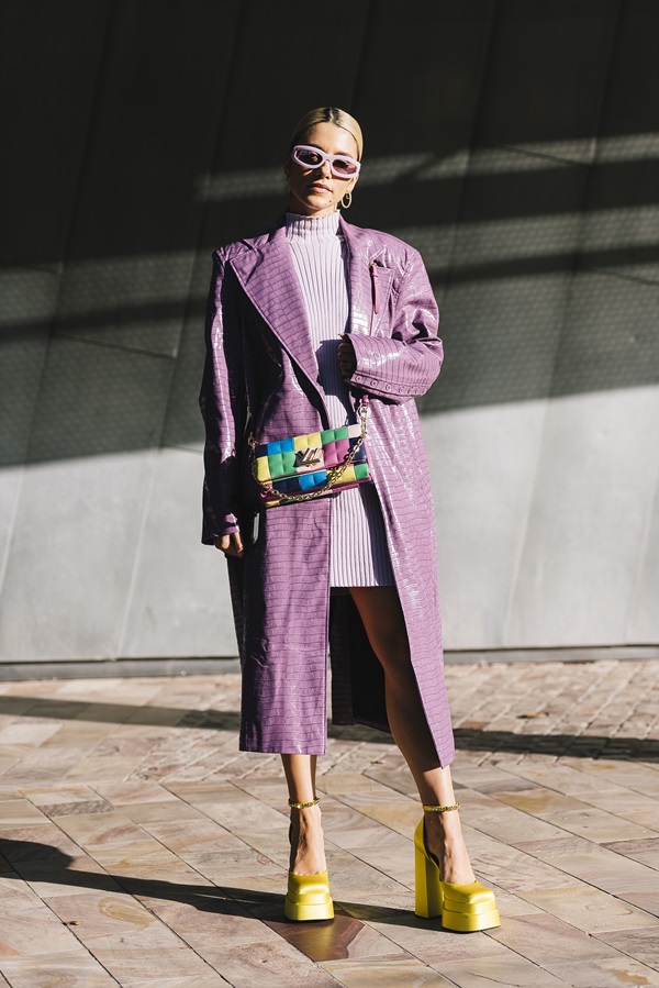 Mulher jovem e branca, com cabelo loiro amarrado, durante a Semana de Moda de Paris, na França. Ela usa um vestido lilás, um casaco de couro roxo e uma sandália de salto alto amarela da Versace. Por cima, usa uma bolsa colorida.