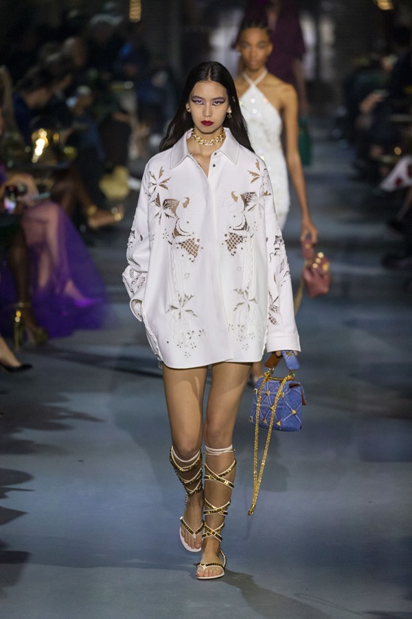 Modelo mulher branca e jovem, com cabelos longos pretos, andando pela passarela em desfile da marca Valentino. Ela usa uma camisa branca de botão com bordados e uma sandália gladiadora, também branca, com taxinhas douradas.