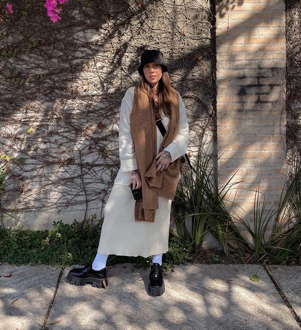 A influenciadora Manoela Meinke, uma mulher jovem, branca e de cabelo liso castanho, posando para foto em um jardim. Ela usa um casaco branco, uma saia de tricô bege, um cachecol marrom, bucket heat preto e um mocassim preto com sola tratorada