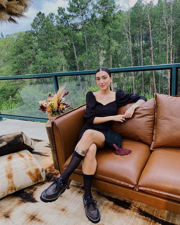 A influenciadora Maju Trindade, uma mulher jovem, branca e de cabelo liso castanho, posando para foto sentada em um sofá de couro marrom. Ela usa um vestido preto curto com mangas bufantes e um mocassim marrom de couro com sola tratorada.