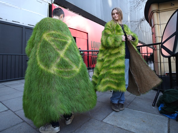 Pessoas brancas e jovens, com cabelo castanho, protestando sobre as mudanças climáticas. Usam um casaco verde com textura de grama para passar a mensagem de necessidade de cuidar do meio ambiente