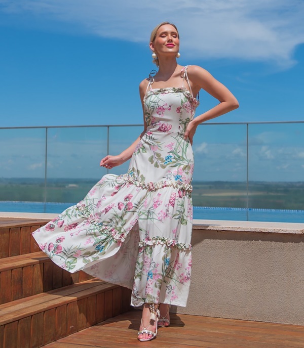 Mulher jovem e branca, com cabelos loiros curtos, posando para foto em um cobertura com uma piscina ao lado. Ela usa um vestido esvoaçante branco com flores rosas e verdes estampadas