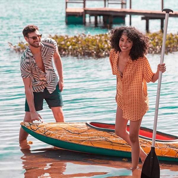 Homem e mulher, ambos jovens, posando para foto na beira do mar, segurando um barquinho e um remo. Ele é branco e tem o cabelo castanho, e usa bermida verde com camisa preta e branca, e a mulher é negra, com cabelo cacheado, usa conjunto de shorts e camisa marrom