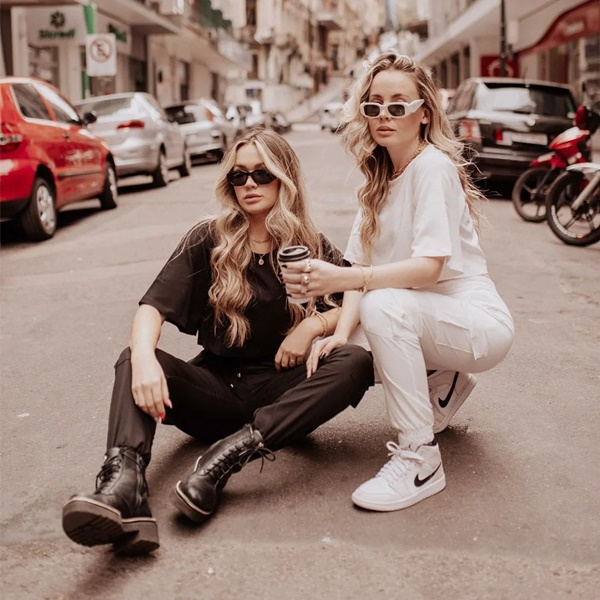 Duas mulheres brancas, jovens e louros, com cabelos longos ondulados, posando para foto no meio da rua. Ambas usam conjuntos de moletom, sendo que uma está com o look todo preto e outra com o look todo branco