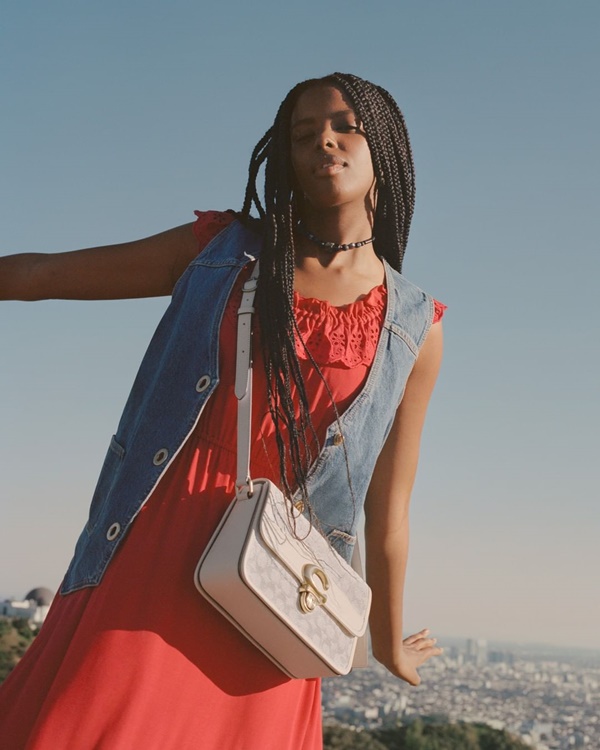 Mulher negra e jovem, com cabelos longos trançados, posando para foto de campanha da marca Coach. Ela usa um vestido vermelho, um colete jeans e uma bolsa branca, tudo da marca