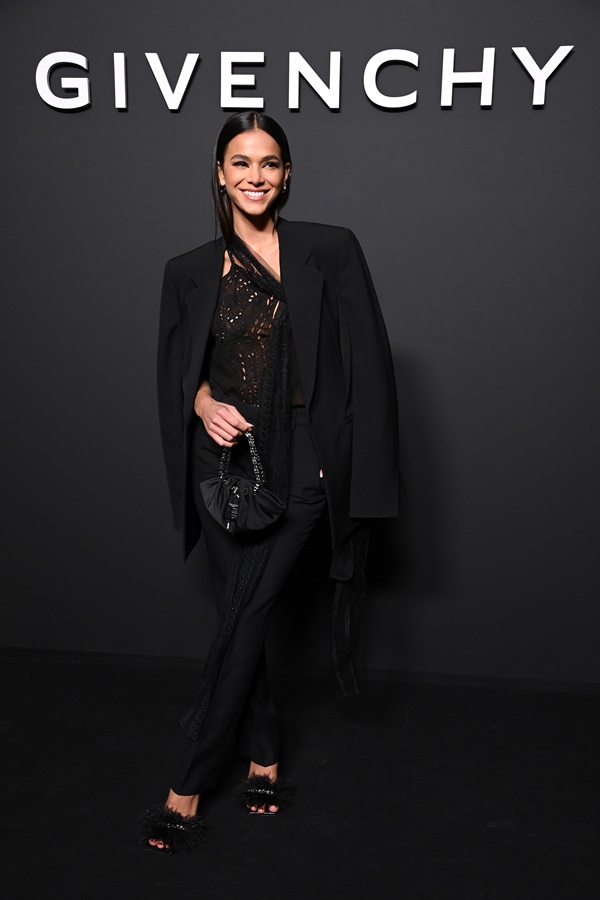 A atriz Bruna Marquezine na porta do desfile da marca Givenchy, na Semana de Moda de Paris. Ela é uma mulher branca e jovem, de cabelos lisos cumpridos, e usa uma blusa de renda, calça reta de alfaiataria e um blazer. Todas as peças são pretas.