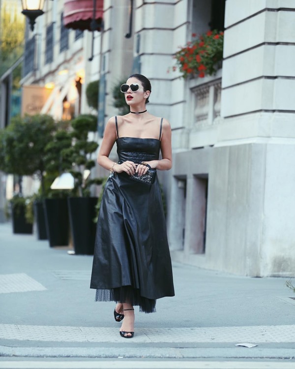 A atriz Bruna Marquezine, uma mulher branca, jovem e com cabelo castanho curto, andando pelas ruas de Paris. Ela usa um vestido longo de couro, uma sandália de salto e uma bolsa. Todas as peças são pretas e da Dior.