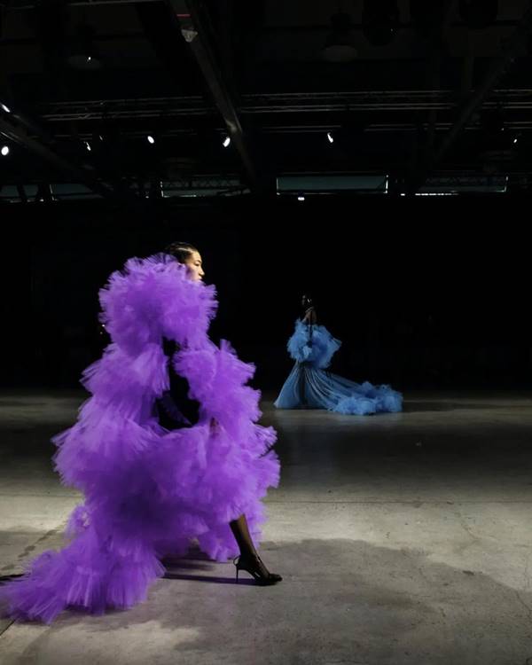 Em desfile, modelo caminha usando look roxo maximalista de tule. Na foto tirada em lateral, é possível ver outra modelo ao fundo com roupa azul, também de tule