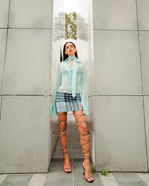 A atriz Bruna Marquezine posando para foto na semana de moda de Paris. Ela é uma mulher jovem, branca e de cabelos curtos e lisos, e usa uma camisa azul transparente, uma saia curta xadrez azul escuro e uma sandália de salto gladiadora com tiras até acima da coxa.