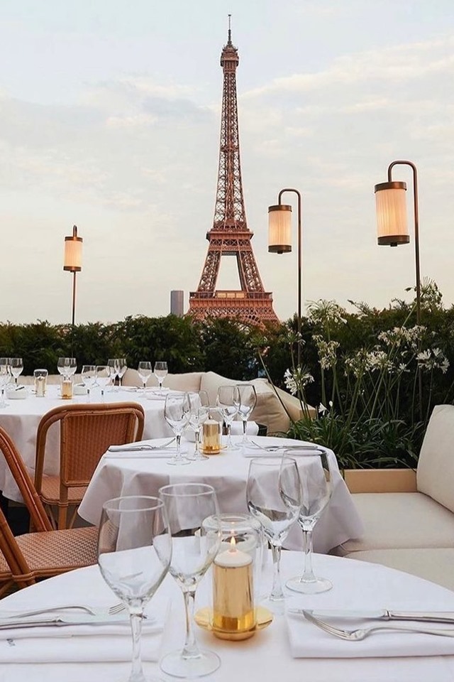 Foto colorida. Ao fundo, aparece a Torre Eiffel. Mesas e cadeiras 