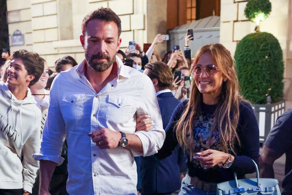 Foto colorida. Homem e mulher de braços dados. Ao redor deles, pessoas tentam tirar fotos