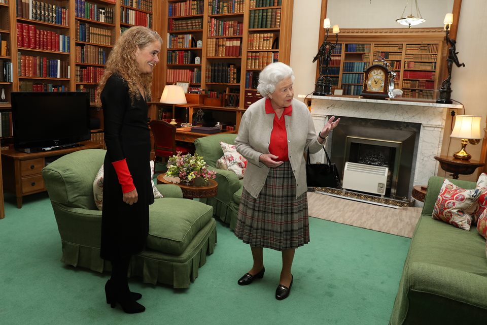 Foto colorida. Duas mulheres conversam em uma sala com estantes cheias de livros e objetos antigos