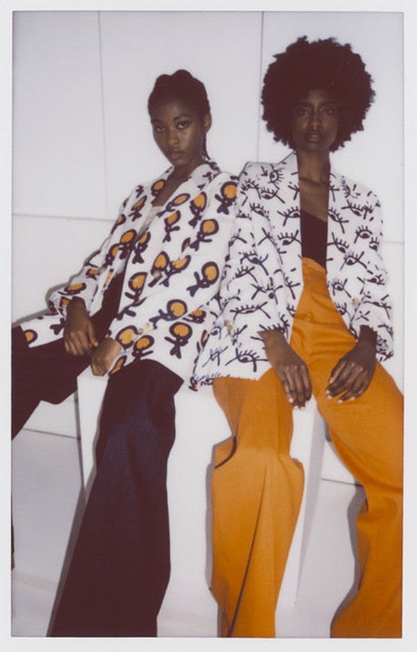 Foto estilo polaroid que divulga a coleção de roupas da marca Asantii. Na imagem é possível ver duas modelso negras e jovens, de cabelos cacheados curtos, calças de alfaiataria azul e mostarda, tops pretos e, por cima, casacos blazers brancos com desenhos em preto e branco