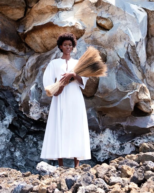 Mulher negra e jovem, de cabelo curto e cacheado, posando para foto em uma praia com rochas. Ela usa um vestido longo branco, com mangas bufantes, e segura um buquê de folhas marrons nas mãos.