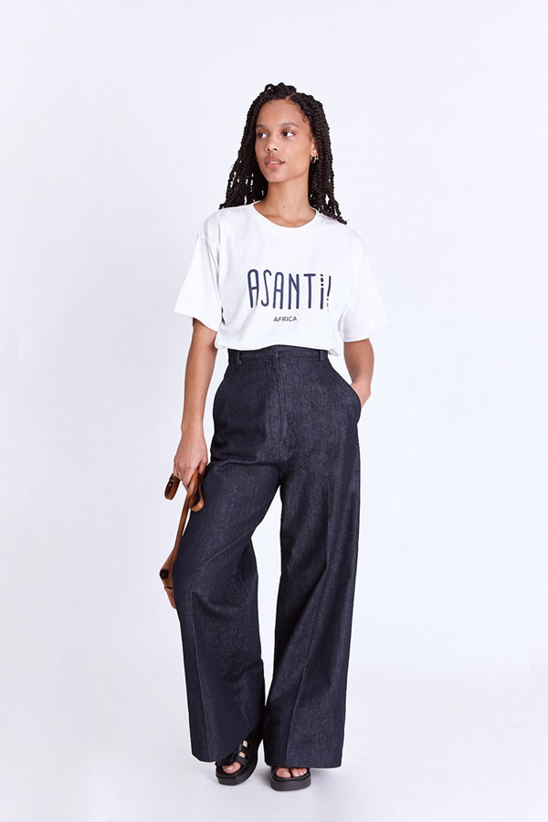 Foto estilo lookbook da marca Asantii, quando a modelo posa com a roupa em um fundo infinito branco. Na imagem é possível ver uma modelo negra e jovem de cabelo comprido trançado usando uma calça azul e uma camiseta branca