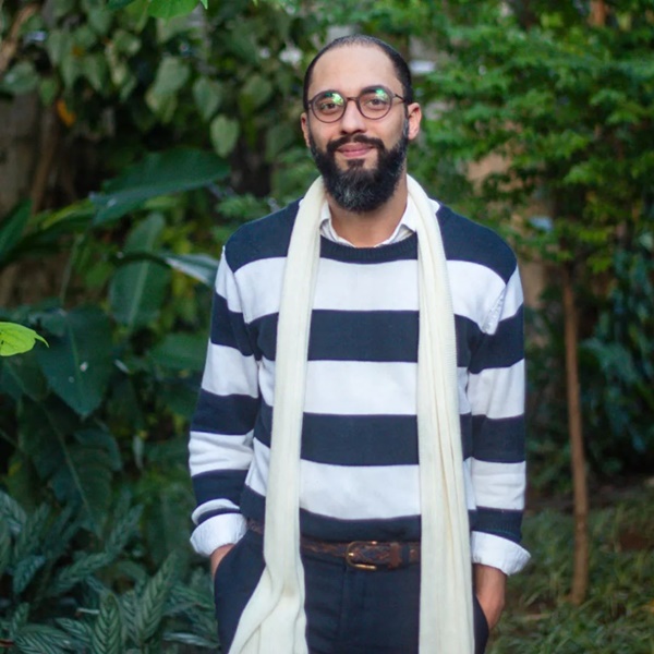 O professor Brunno Almeida Maia, um homem branco e jovem, de barba e cabelo preto, posando para foto em um jardim. Ele usa um casaco listrado branco e azul e um cachecol branco