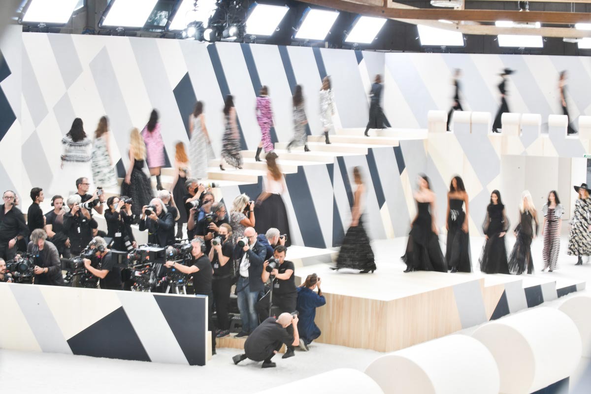 Desfile da marca Chanel. Na foto é possível ver uma passarela com modelos desfilando e fotógrafos na ponta do palco