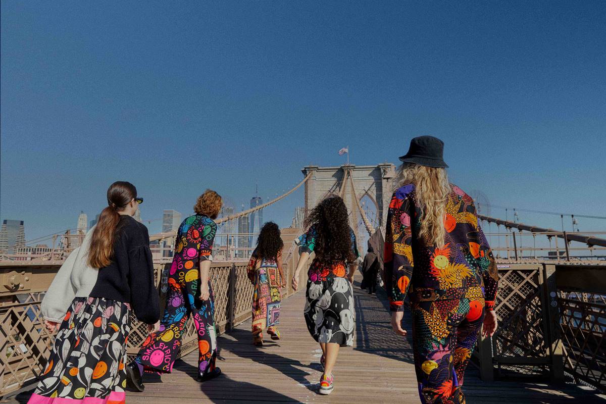 Pessoas usando roupas da marca carioca FARM e andando pela Ponte do Brooklyn, em Nova York, nos Estados Unidos. Elas usam roupas pretas com estampas de emojis sorrindo, todos coloridosv