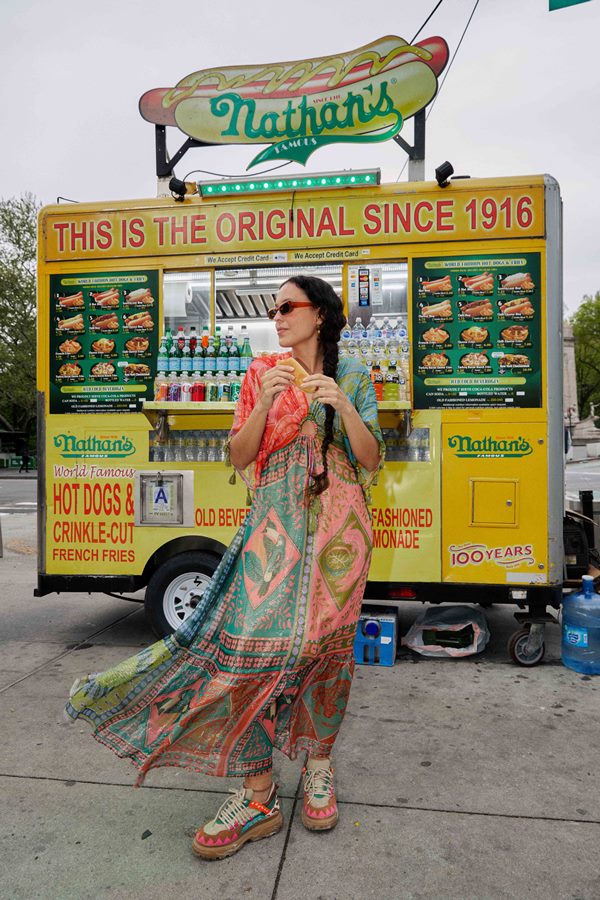 Mulher branca e jovem, com cabelo liso e longo, posando para foto em frente a um foodtruck em Nova York, nos Estados Unidos. Ela usa um vestido estampado verde, rosa e vermelho da marca FARM e óculos escuros.