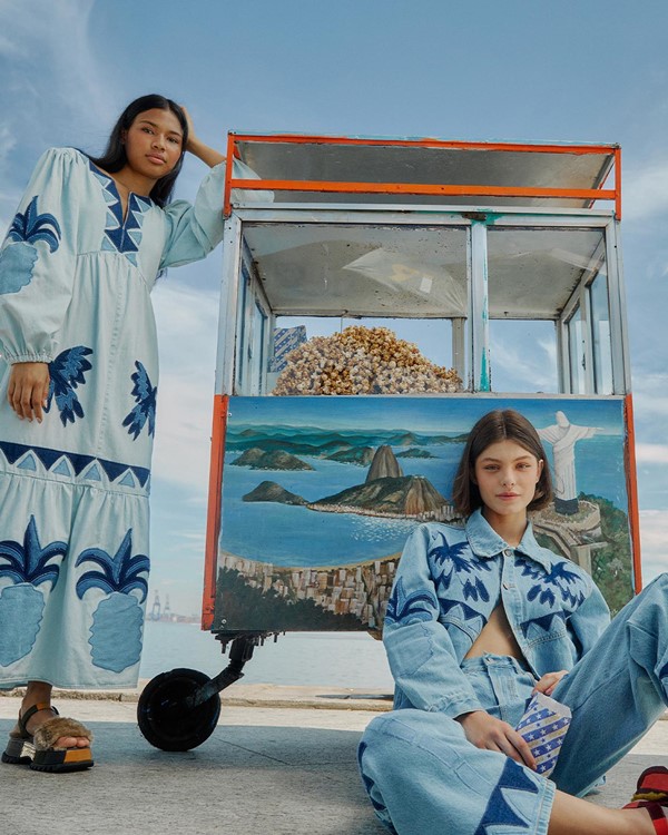 Duas mulheres posando para foto em um carrinho de pipoca na orla da praia do Rio de Janeiro. A foto é de uma campanha da FARM e é possível ver uma modelo jovem e indígena com um vestido azul claro e outra modelo jovem, branca, e de cabelo liso vestindo uma camisa e uma calça jeans.