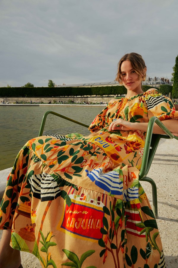 Modelo branca e jovem, de cabelos curtos lisos, sentada na beira do Rio Sena, em Paris, na França. Ela usa um vestido amarelo com estampas de flores, guarda-sóis e o refrigerante cajuína