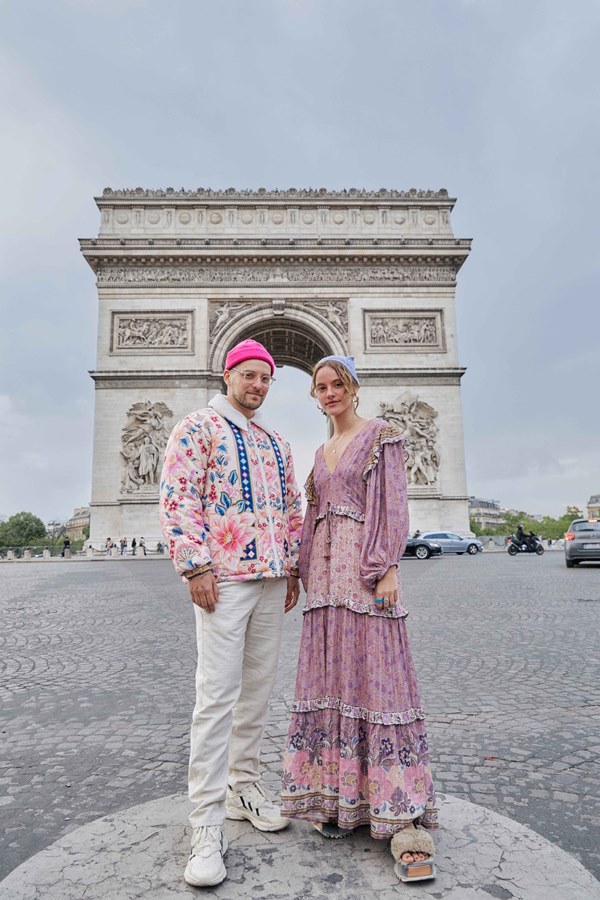 Homem e mulher, ambos brancos e jovens, posando para foto em frente ao Arco do Triunfo, em Paris, na França. Ele usa uma calça branca, um casaco rosa estampado e uma touca rosa neon; já ela usa um vestido longo com estampa rosa de florzinhas. A foto é da campanha da coleção da marca FARM.