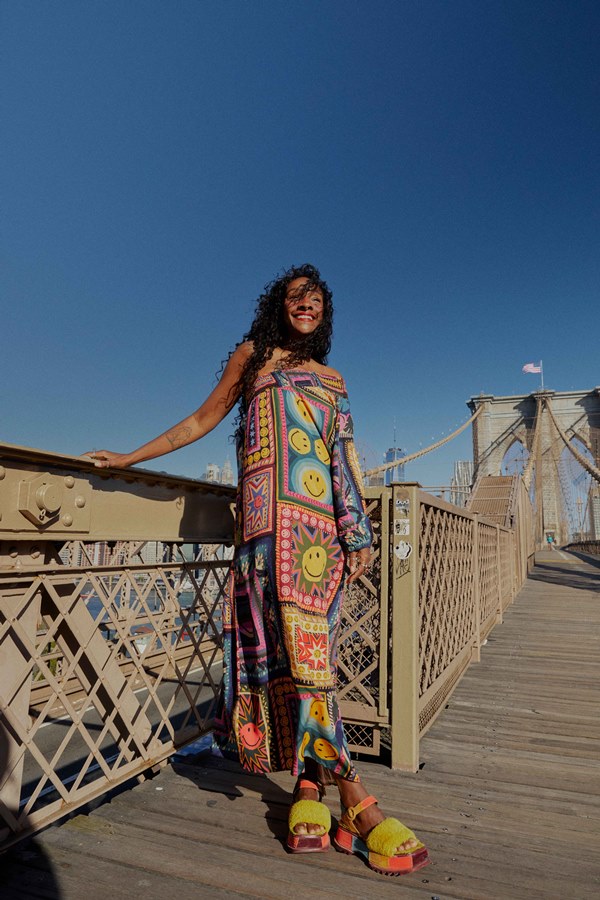 Mulher jovem e morena, de cabelo cacheado longo, posando para foto na Ponte do Brooklyn, em Nova York, nos Estados Unidos. Ela usa um vestido longo com estampa de emojis sorrindo coloridos.