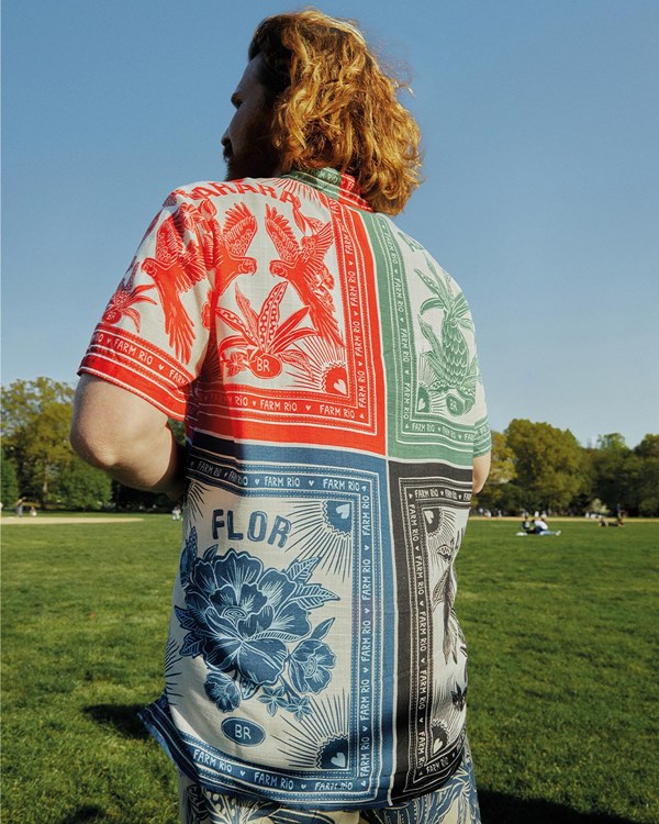 Homem branco e jovem, de cabelo e barba ruiva, posando para foto em um jardim. Ele usa uma camisa de botão estampada, com um quadrado vermelho, um azul, um verde e outro preto. A roupa é da marca FARM.